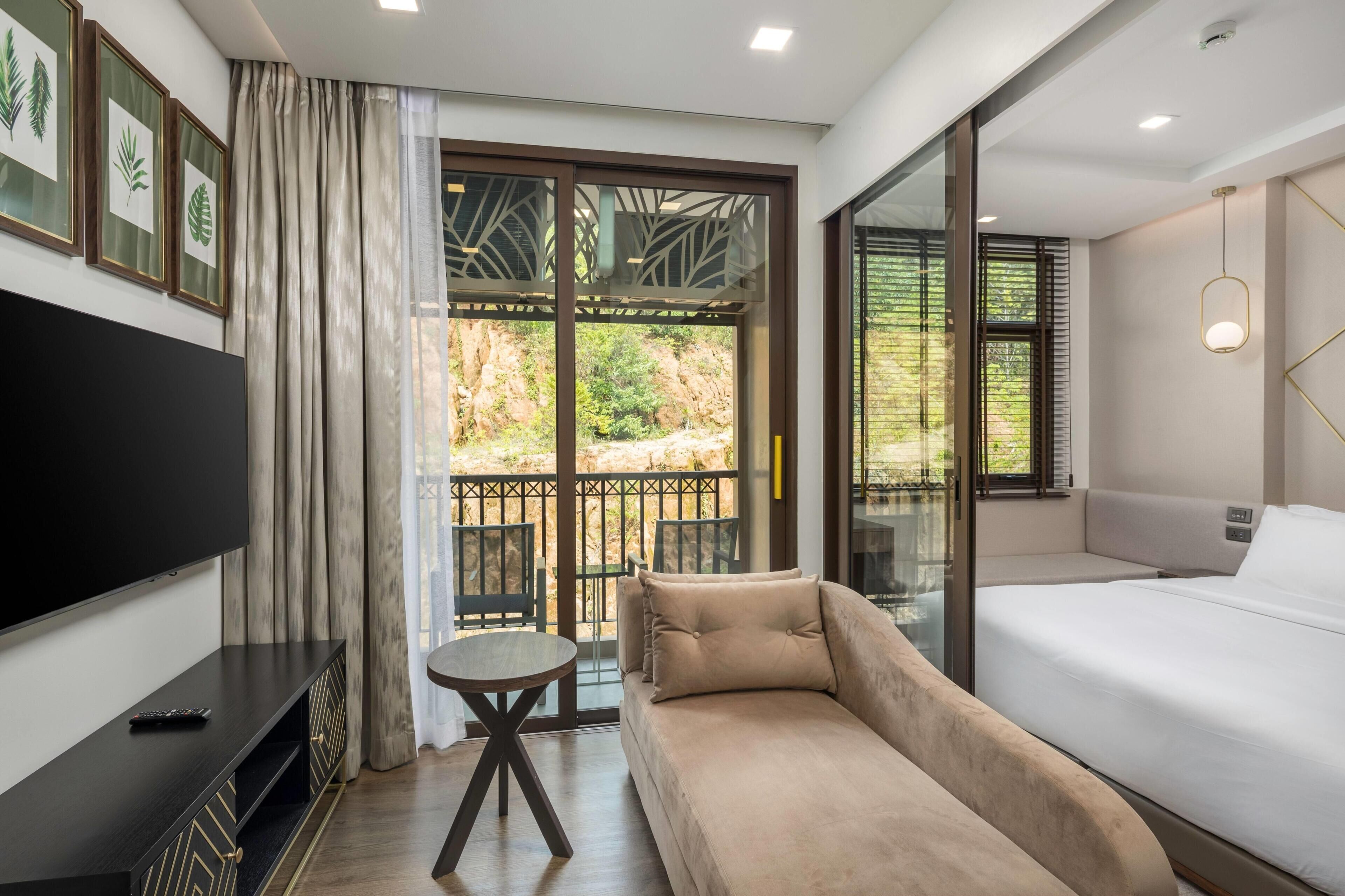 Una habitación de hotel moderna con una cama grande, un televisor, una mesita y un balcón con vistas a zonas verdes.