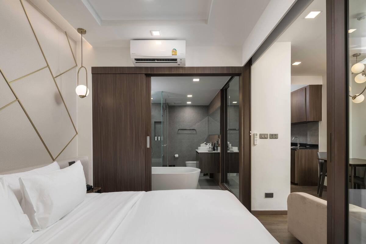 Une chambre d’hôtel moderne équipée d’un lit, d’un appareil de climatisation, d’une salle de bain fermée par des parois de verre et d’une kitchenette.