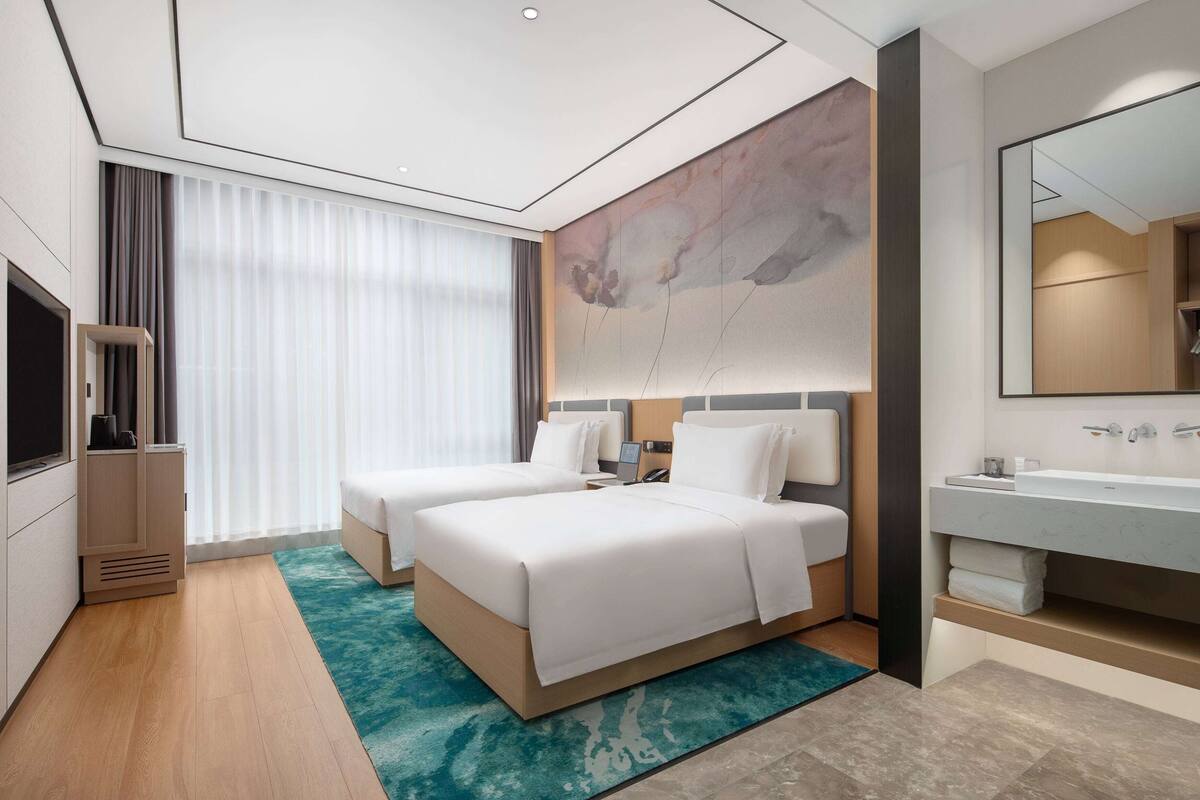 Une chambre d’hôtel moderne avec deux lits, un grand miroir, une salle de bain avec un lavabo et des toilettes, et une grande fenêtre avec des rideaux.