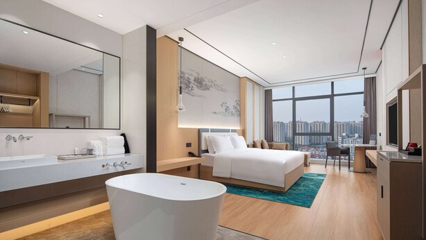 A modern hotel room with a large bed, a separate bathroom with a bathtub, a large window offering a city view, and a desk with a chair.