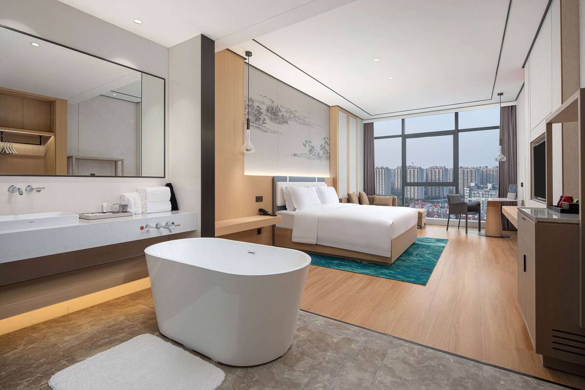 Une chambre d’hôtel moderne dotée d’un grand lit, d’une salle de bain séparée avec baignoire, d’une grande fenêtre donnant sur la ville et d’un bureau avec une chaise.