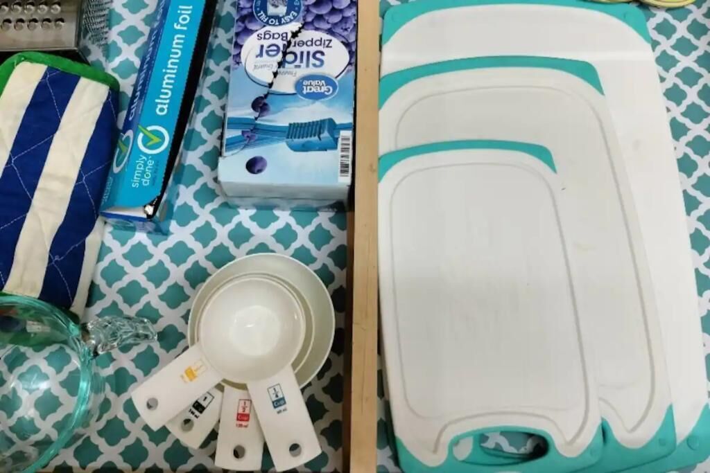 Una encimera de cocina con un electrodoméstico blanco y aguamarina, tazas medidoras, una caja de papel de aluminio y un paño a rayas.