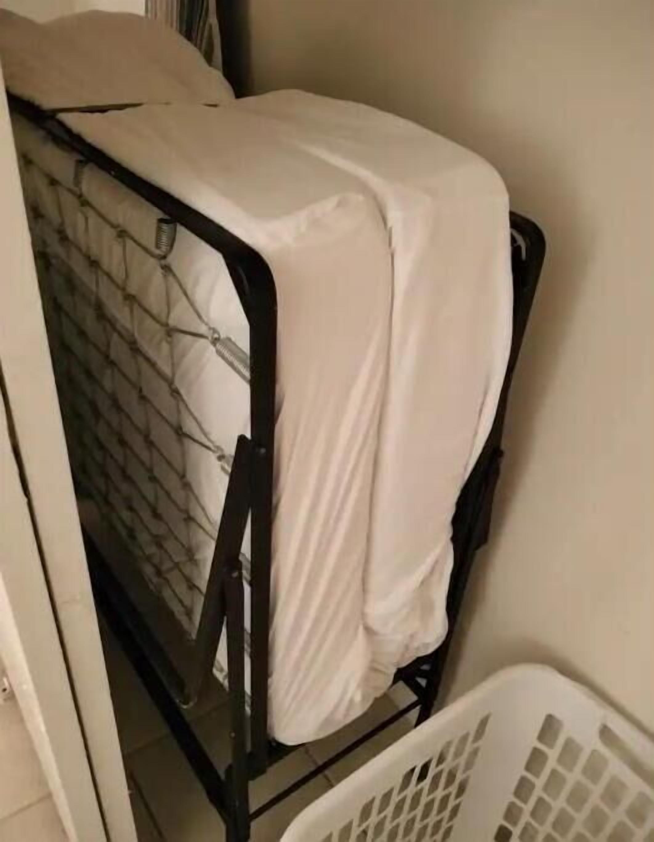 A small, enclosed space with a metal bed frame and a white mattress. A white laundry basket is visible in the foreground.