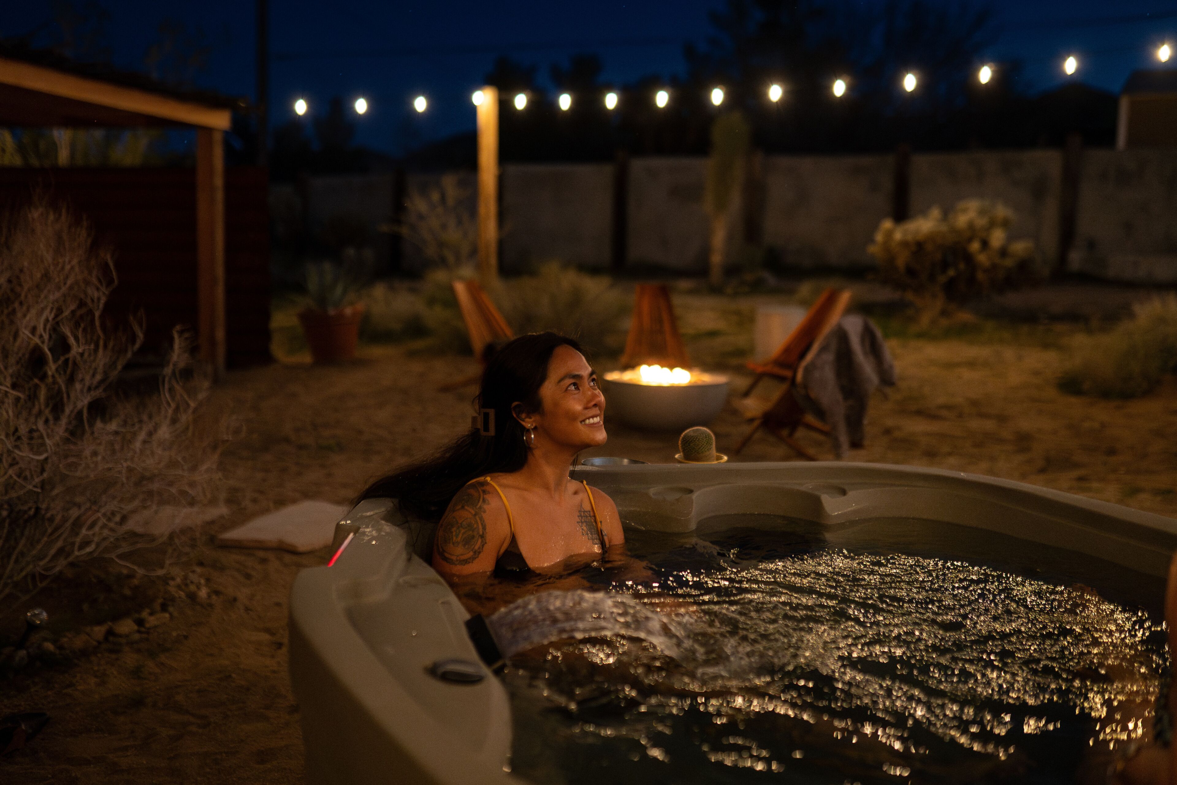 A person relaxing in a hot tub at night with string lights in the background.
