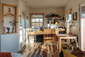 A compact kitchen with wooden cabinets, a dining area with a table and chairs, a dog sitting by the window, and a variety of kitchen utensils and appliances.