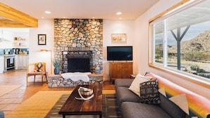 A living room with a stone fireplace, a sofa, a coffee table, and a TV. - Top 1% | 4min->JTNP+Town|Cinema|Hot Tub|Fire Pit (Joshua Tree)