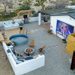 A backyard with a hot tub, a fire pit, outdoor seating, and a TV screen.