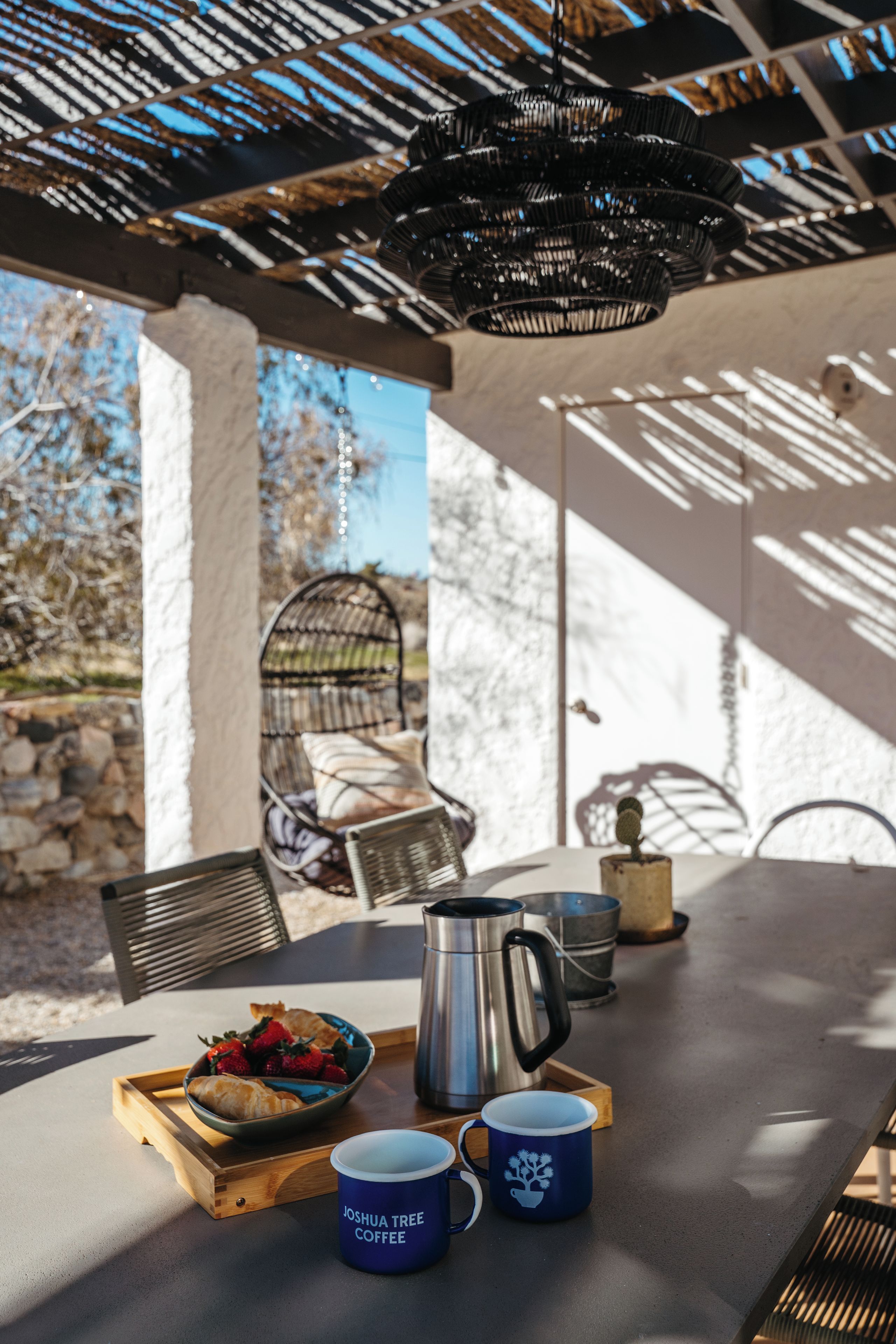 Un área de comedor al aire libre con una mesa dispuesta para dos, que incluye una jarra de metal, una taza azul y una bandeja de madera con comida.
