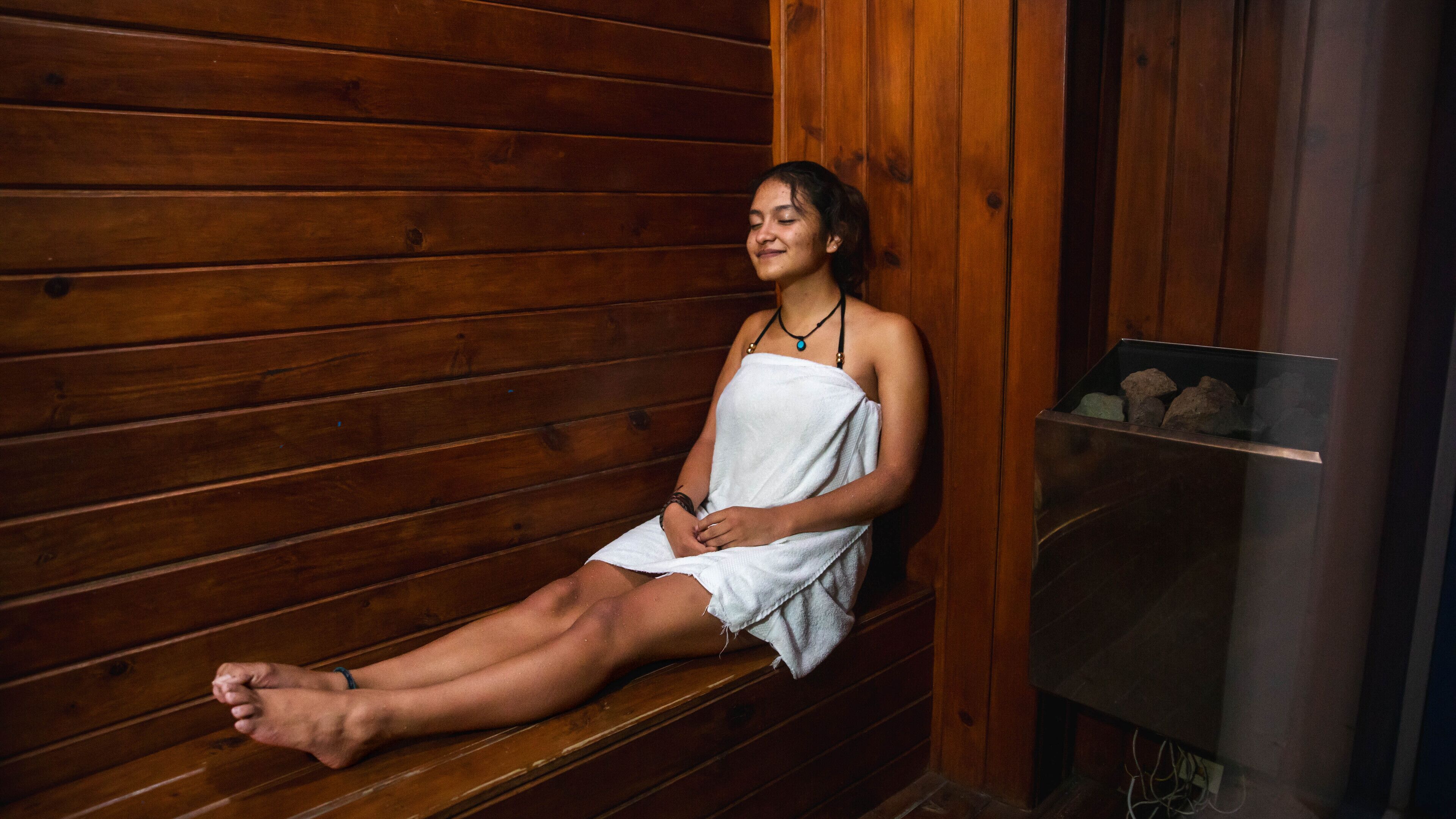 A person sitting on a wooden bench in a sauna room with a stone wall and a small shelf with rocks.