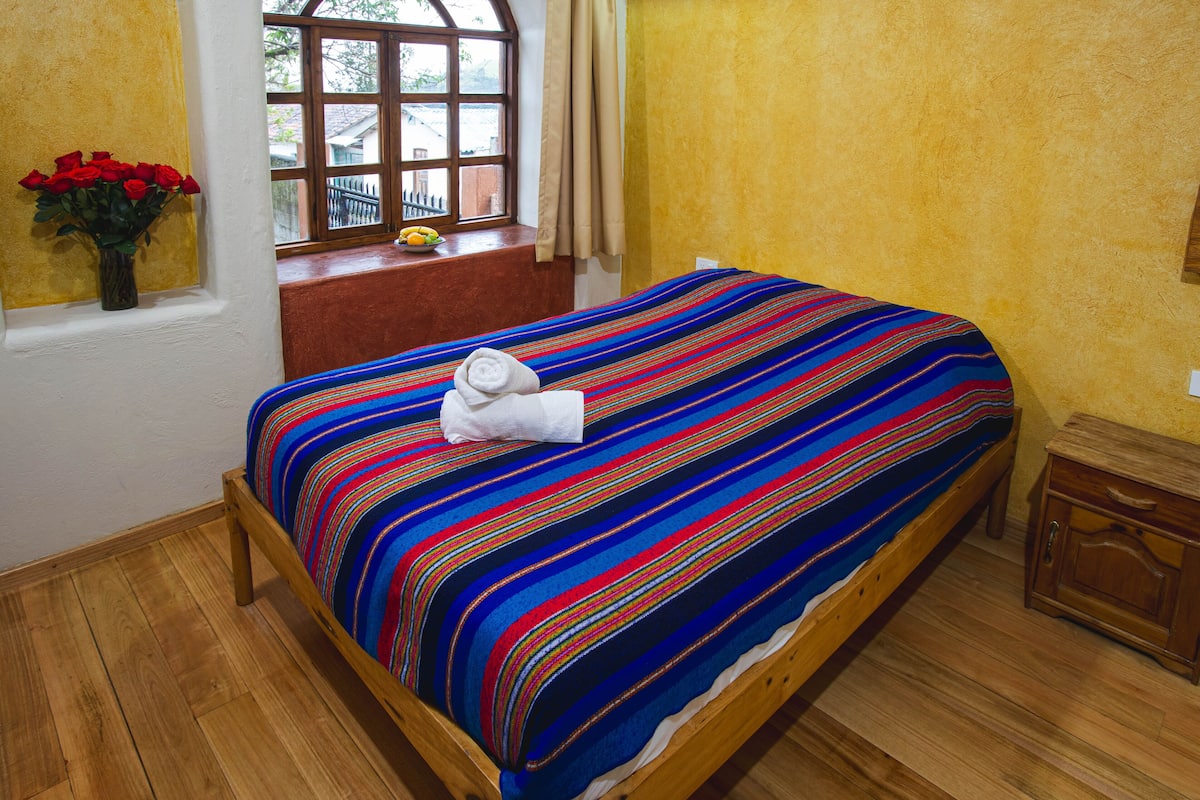 Una cama con una colcha a rayas de colores, una ventana que da a una valla, y un florero con flores rojas en el alféizar.