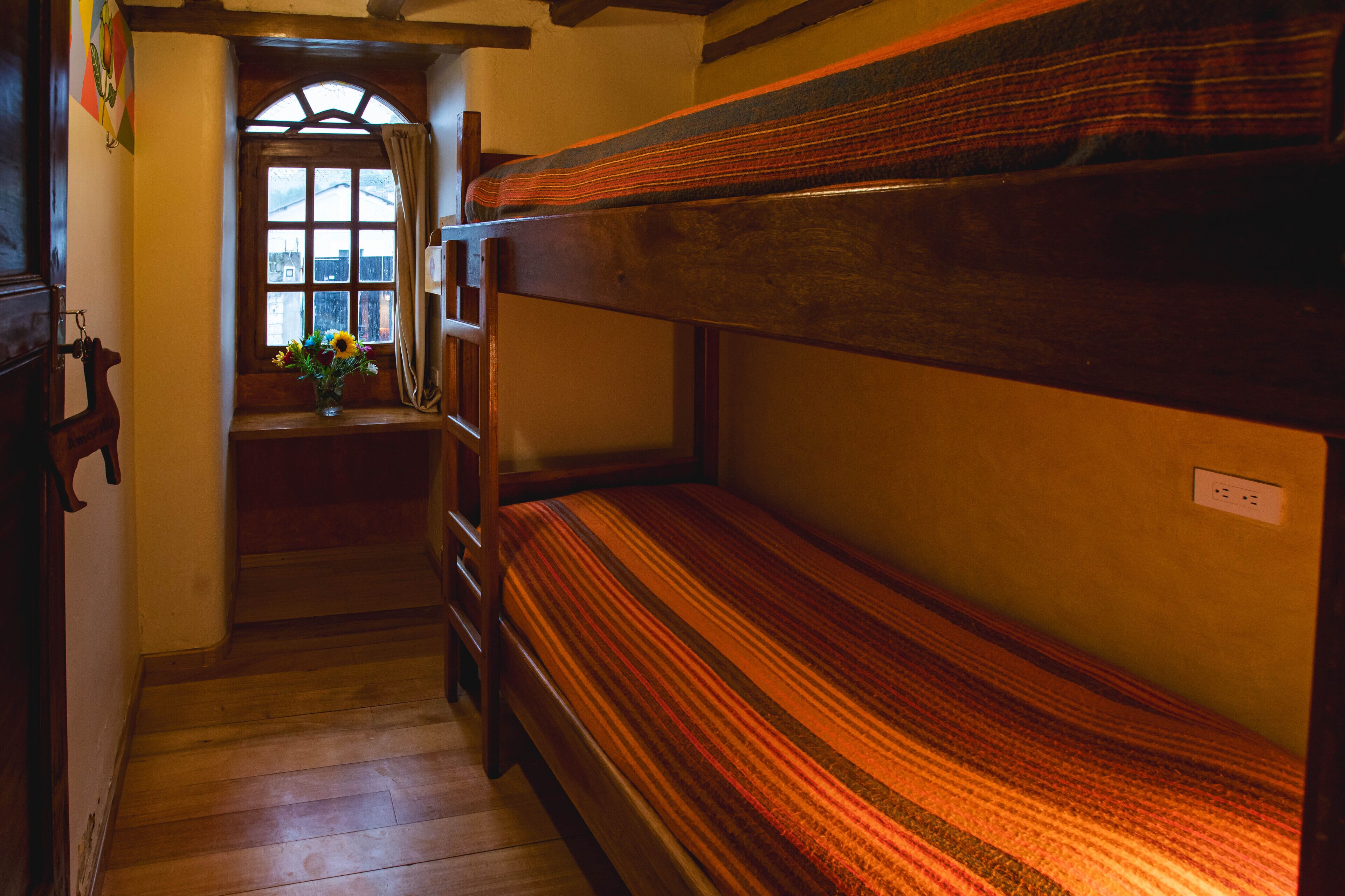 Een slaapkamer met stapelbedden, een houten vloer, een raam met gordijnen en een klein tafeltje met een bloemenvaas.