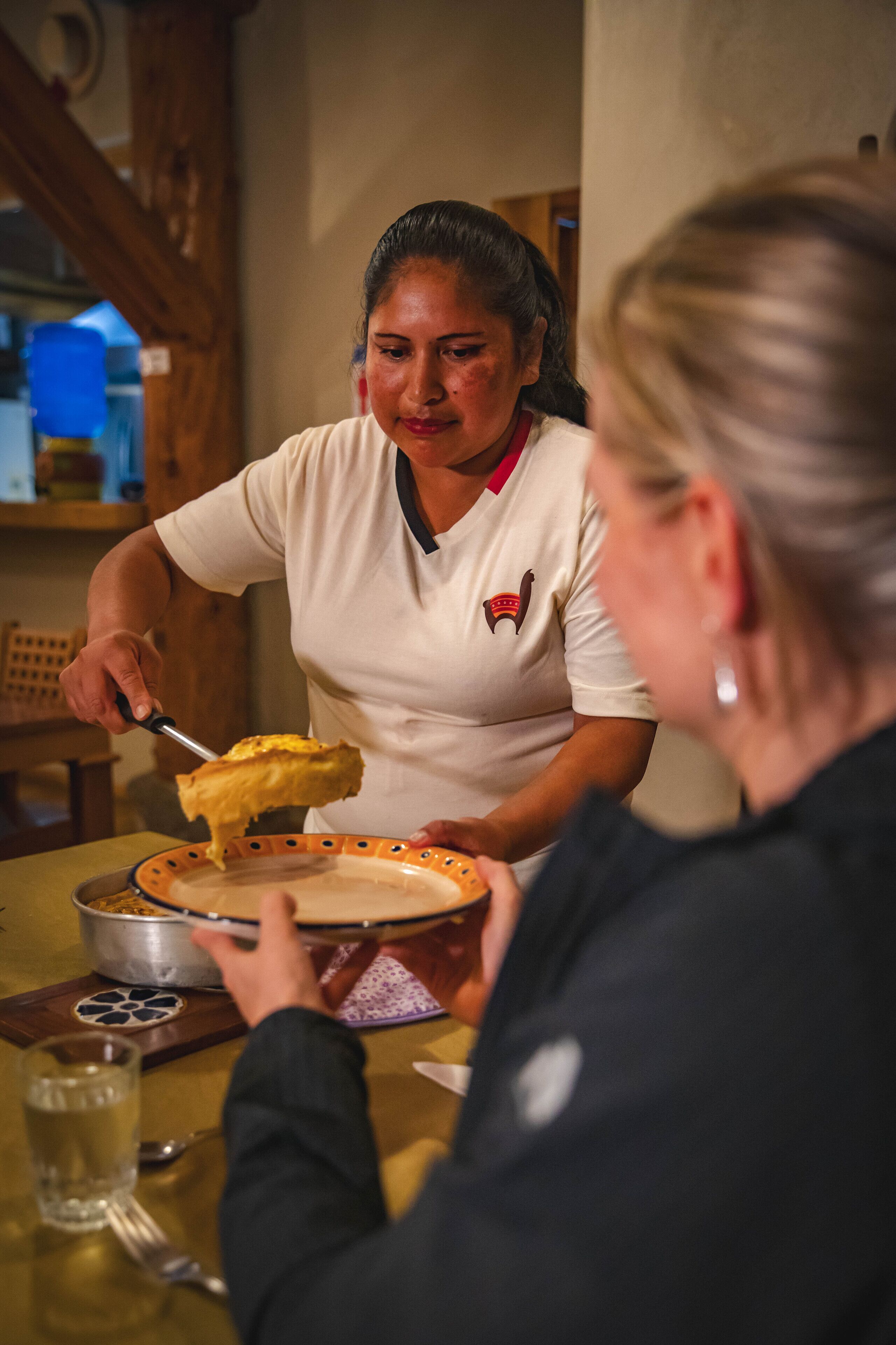 Una persona sirve tarta a otra sentada a una mesa, con un vaso de agua y una cuchara.