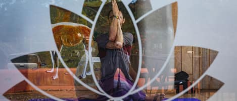 Una persona practicando yoga sobre una colchoneta en una habitación con suelo de madera y una ventana que da a vistas de árboles.