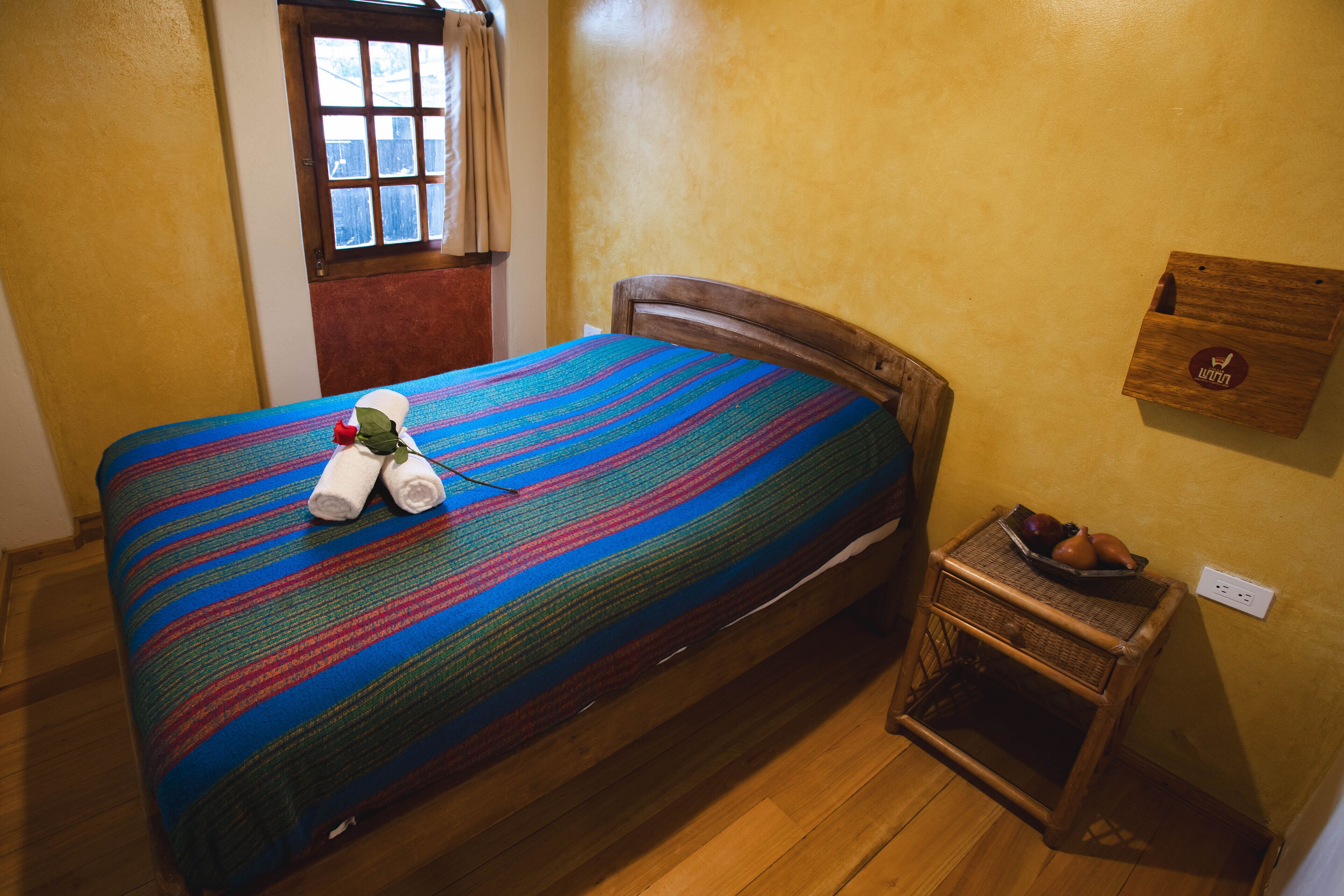Un letto con una coperta a righe colorate, un piccolo comodino di legno con una ciotola di frutta e una finestra con tende.