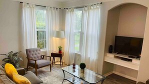 Ein Wohnzimmer mit Sofa, Sesseln, einem Glastisch und einem Fernseher auf einem Schrank.