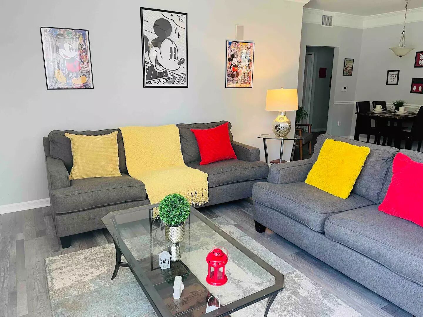 A living room with two grey sofas, yellow and red throw pillows, a glass coffee table with a plant and a red lantern, and framed artwork on the wall.