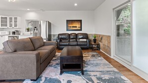 A modern living room with a sofa, a coffee table, and a fireplace. There is a kitchen in the background with white cabinets and a refrigerator. - Private Mountainside Home - close to hiking trails and rivers. (Chilliwack)