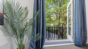 A room with a potted plant, a door leading to a balcony with a black railing, and a view of trees and a road. - Private Mountainside Home - close to hiking trails and rivers. (Chilliwack)