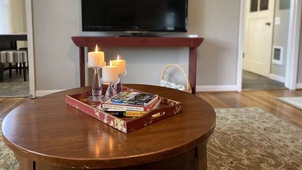 A round wooden table with a tray holding books and candles, a flat-screen TV mounted on a wall, and a door in the background. - Cozy 2 br. Close to River (Milwaukie)