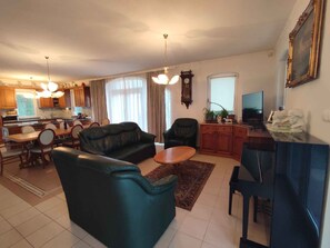 Un salon avec un canapé, des fauteuils, une table basse, un téléviseur et, au fond, une cuisine.