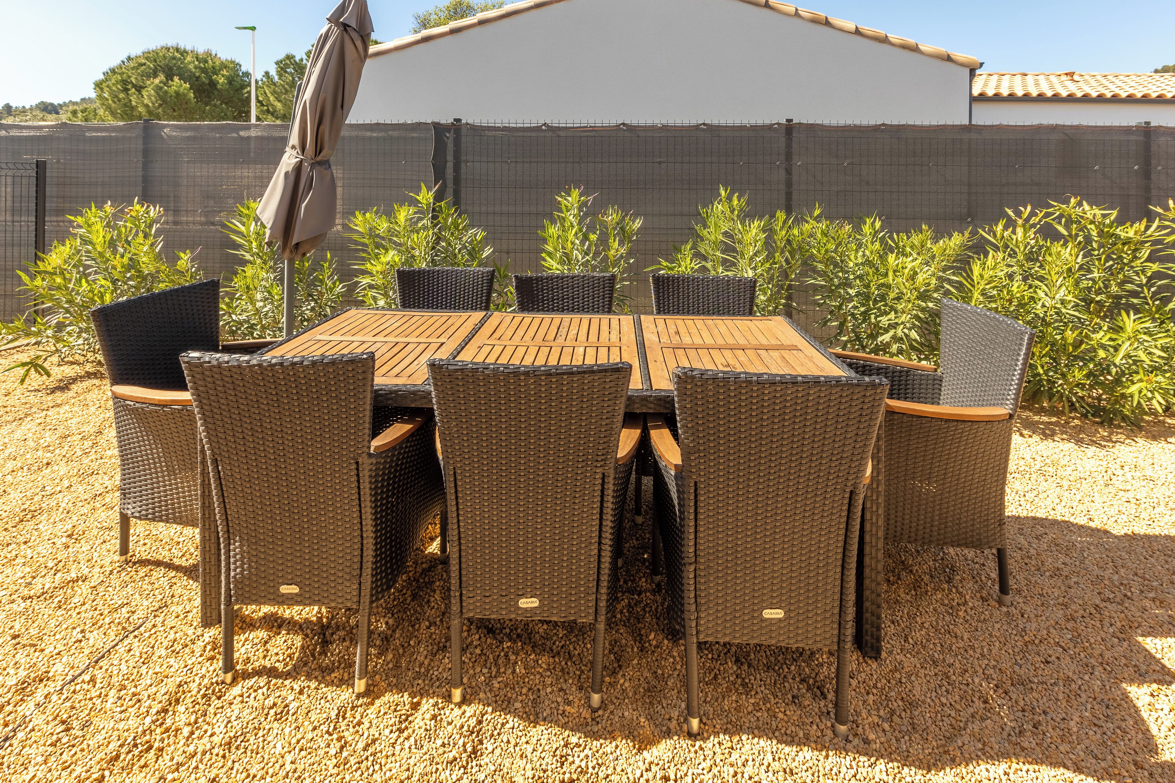 An outdoor patio with a wooden table and wicker chairs, surrounded by gravel and plants.