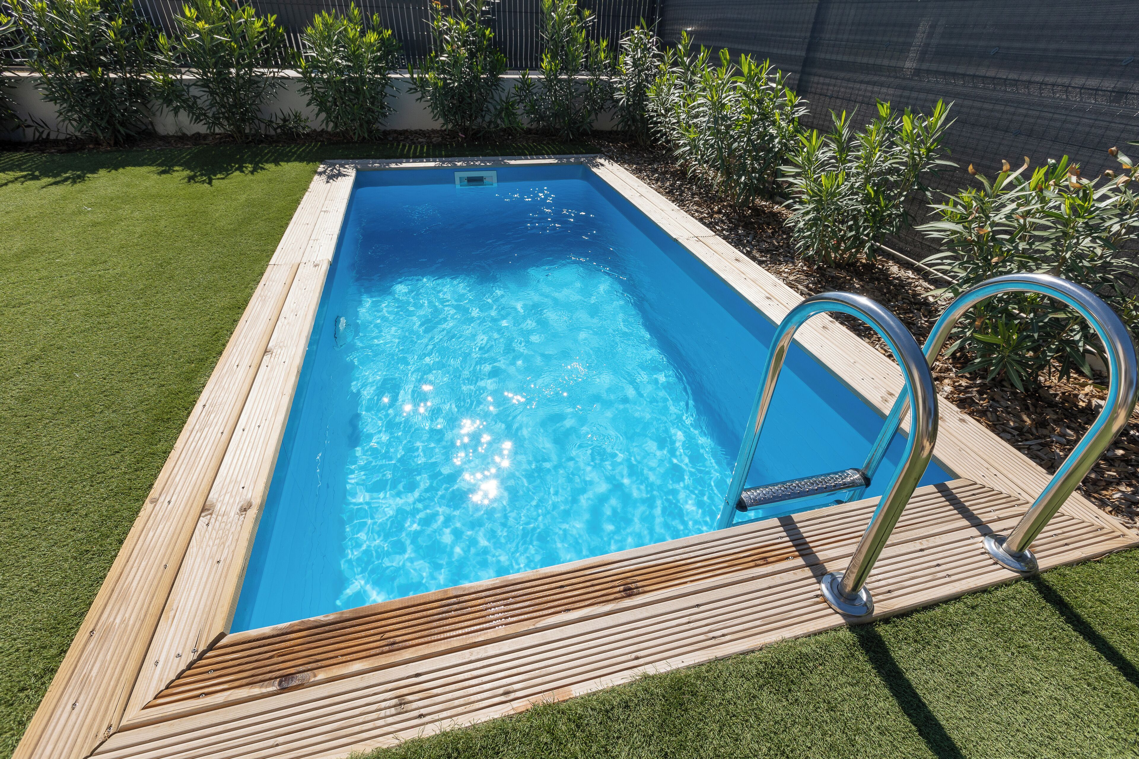 A rectangular swimming pool with a wooden deck and metal ladder, surrounded by a well-maintained lawn and various plants.
