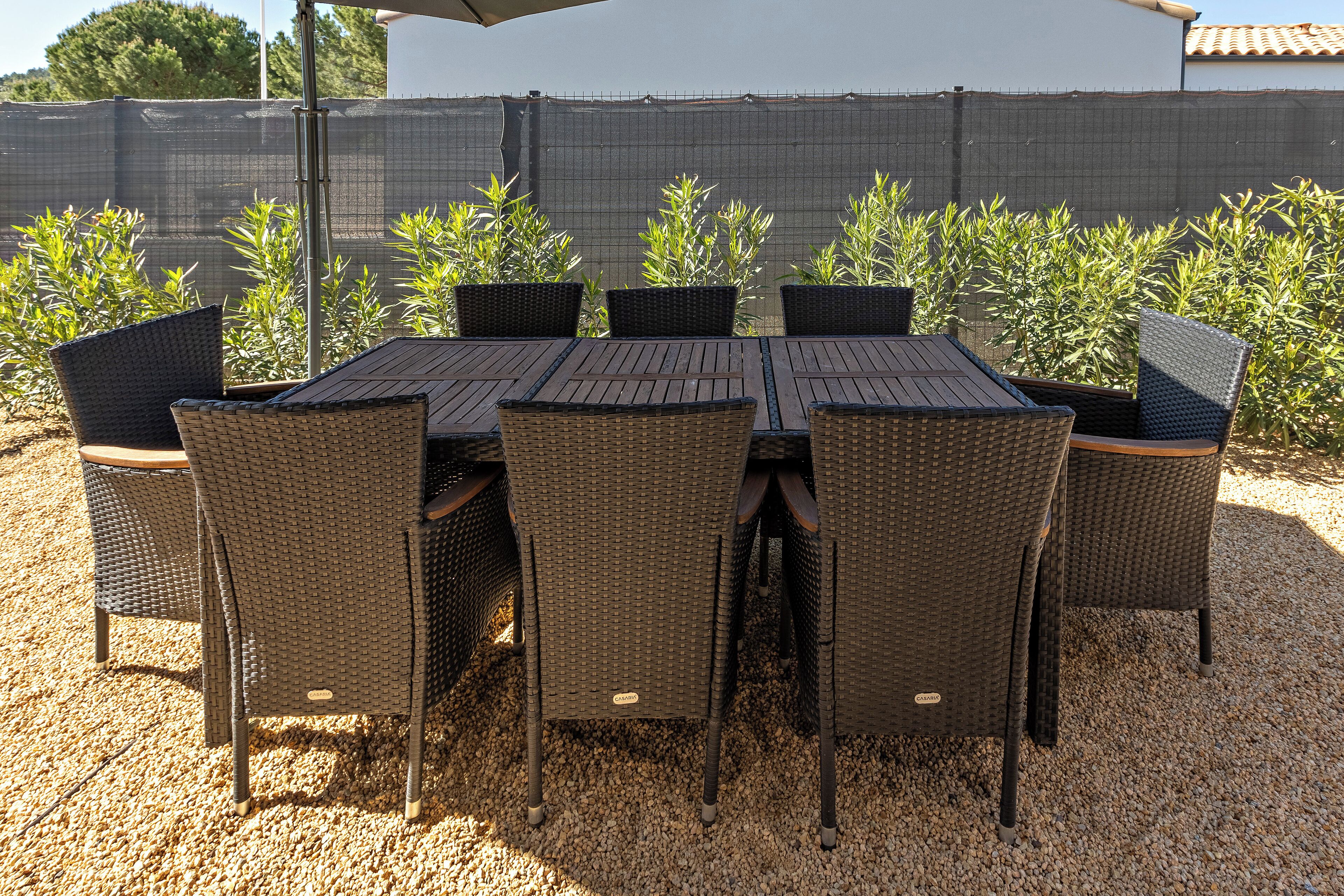 An outdoor patio with a table and chairs, surrounded by a gravel ground and a fence with plants.