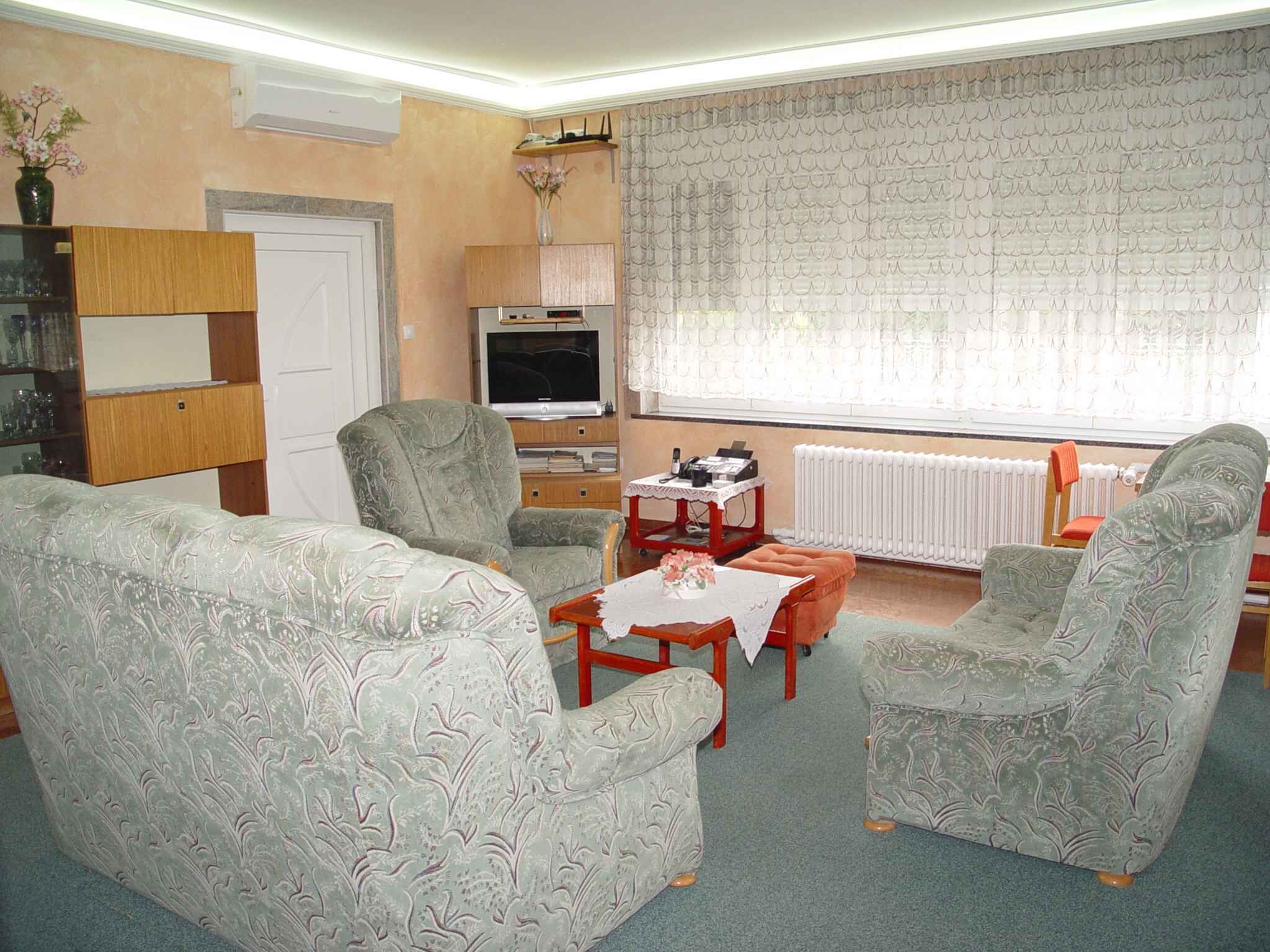 Un salón con sillones estampados, un televisor, un radiador y una pequeña mesa con un florero.