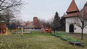 Une aire de jeux dotée de diverses structures ludiques, d’une zone herbeuse et d’un bâtiment avec un toit recouvert de tuiles rouges, visible au fond.