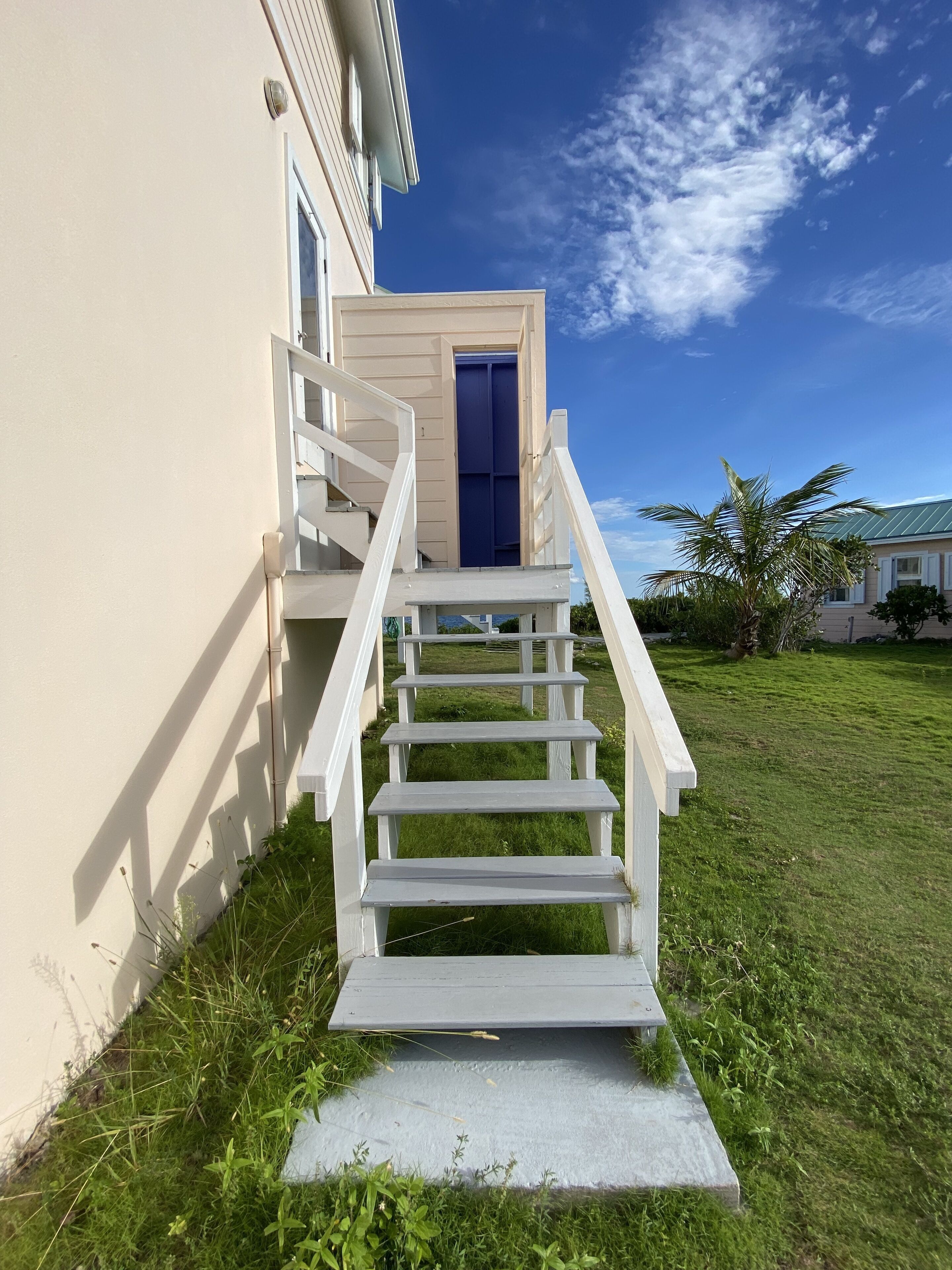 A white staircase leading to a blue door, surrounded by a grassy lawn and a clear blue sky.