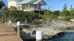 A wooden pier leading to a house with a green roof, surrounded by vegetation and a rocky shoreline. - Private Oceanfront Home on the Bay of Great Guana Cay - Spectacular Views! (Great Guana Cay)