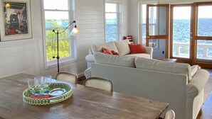 A dining area with a wooden table, chairs, and a green tray with glassware. There is a white sofa with red pillows, a ceiling fan, and windows with a view of the ocean. - Private Oceanfront Home on the Bay of Great Guana Cay - Spectacular Views! (Great Guana Cay)
