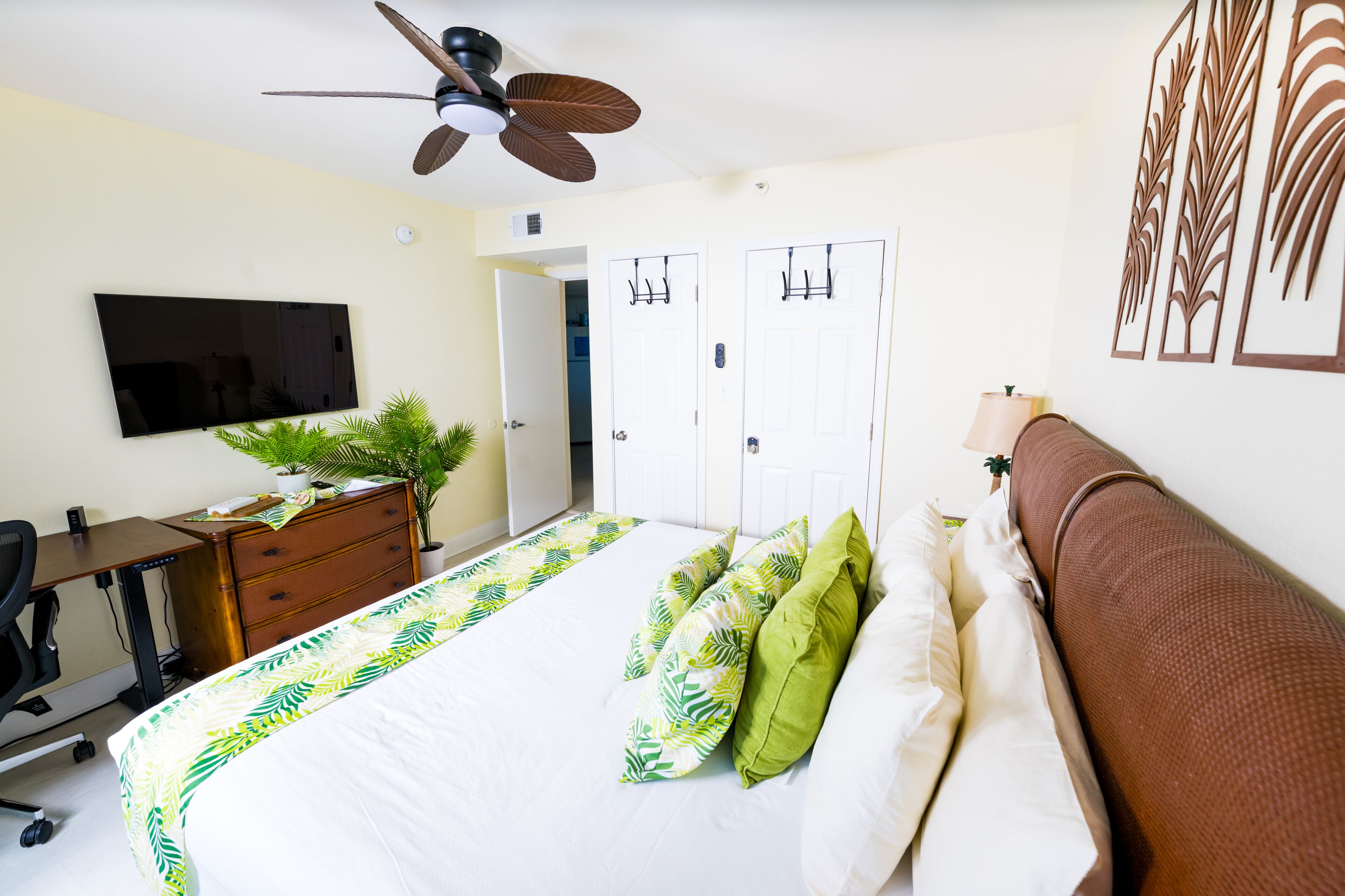 Ein Schlafzimmer mit Bett, Schreibtisch, Stuhl, Fernseher, Deckenventilator und Tür.