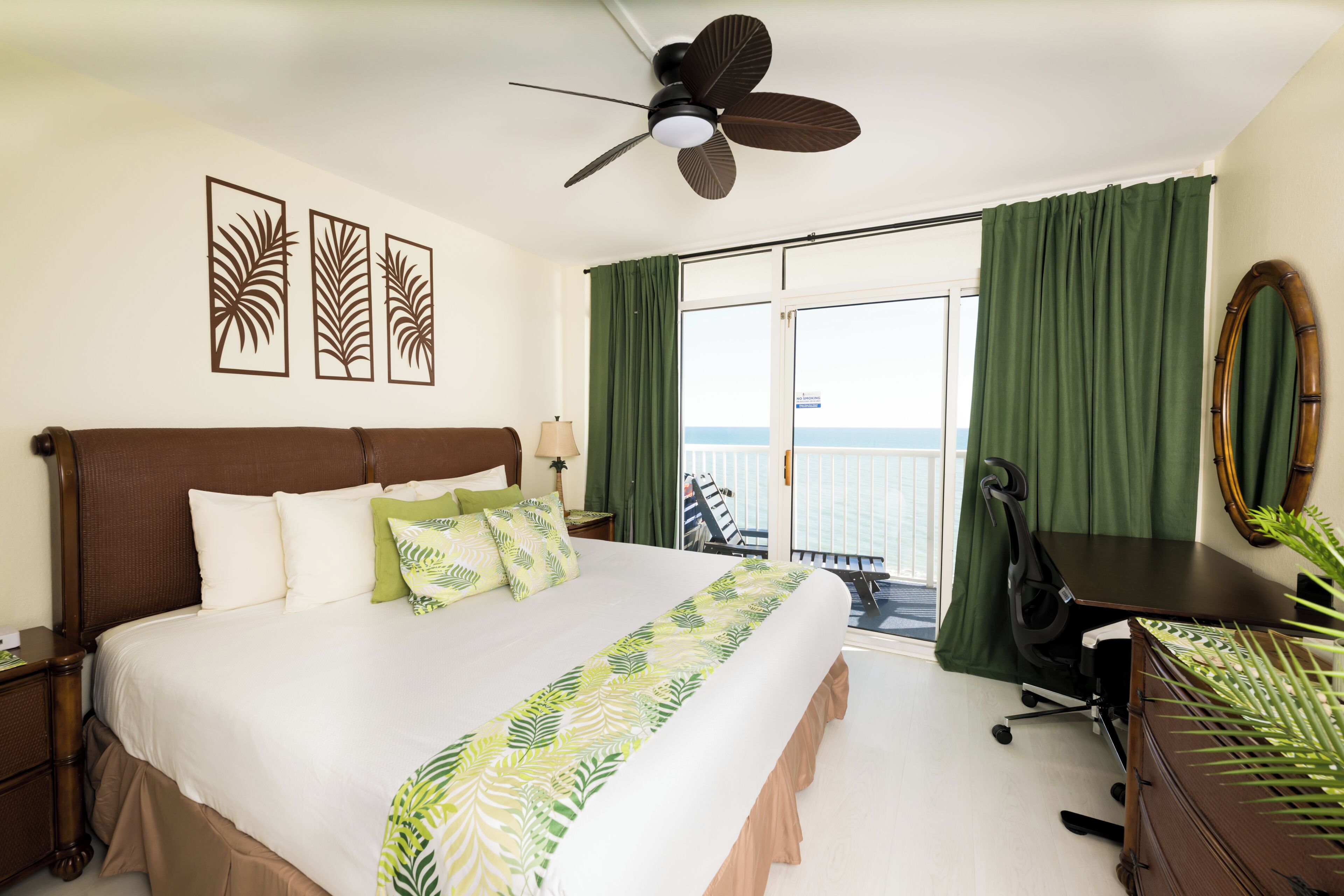 Une chambre d’hôtel équipée d’un lit, d’un bureau, d’une chaise, d’un ventilateur de plafond, d’une grande fenêtre avec des rideaux et offrant une vue sur la mer.