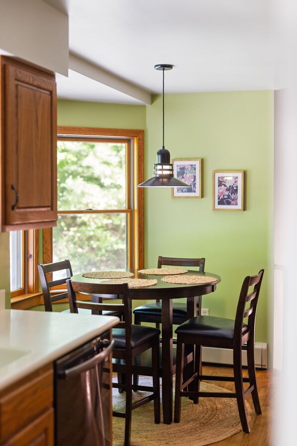A dining area with a round table, four chairs, a hanging pendant light, and two framed pictures on the wall.