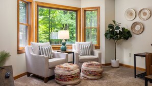 Ein gemütliches Wohnzimmer mit zwei Sesseln, einem Couchtisch und einer Stehlampe. Große Fenster bieten Blick auf einen Wald.