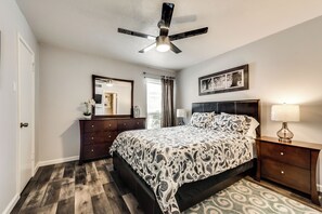 Quarto com cama, cômoda, mesa de cabeceira, ventilador de teto, janela com cortinas e um quadro na parede.