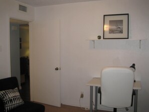Um quarto com porta fechada, uma pequena mesa com uma cadeira, um quadro na parede e uma almofada com padrão preto e branco sobre a cadeira.