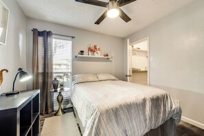 A bedroom with a bed, a desk with a lamp, a ceiling fan, a window with curtains, and a shelf with decorative items. - I00 - Gated Irving 2BR | DFW 10Min | Patio + Pool (Irving)