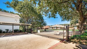 A gated parking area with a covered walkway, a building with a number sign, and trees in the background. - I00 - Gated Irving 2BR | DFW 10Min | Patio + Pool (Irving)