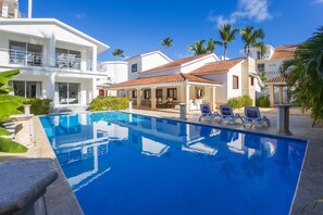 A swimming pool with lounge chairs, a white building with balconies, and palm trees. - Beachfront 8BR Villa with Pool & Maid (Punta Cana)
