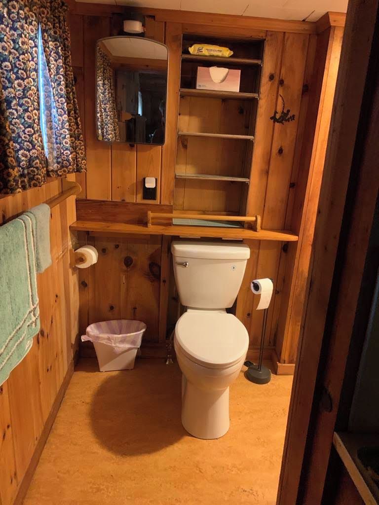 Een kleine houten badkamer met toilet, een klein prullenbakje en een plank met spiegel en toiletpapier.