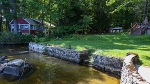 A house with a red exterior and white trim, surrounded by lush greenery and a small stream. - Quiet Brandy Pond Retreat with Sunset Views & Water Access (Naples)