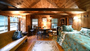 A cozy cabin interior with a wooden ceiling, a round dining table surrounded by chairs, a sofa, and a bed with floral bedding. - Quiet Brandy Pond Retreat with Sunset Views & Water Access (Naples)