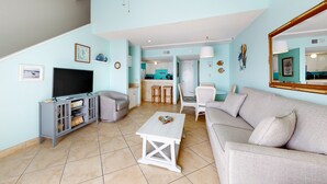 A living room with a TV, sofa, coffee table, and a kitchen in the background. - Sugar Beach Condominiums (Orange Beach)