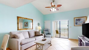 A living room with a sofa, a coffee table, a ceiling fan, a TV stand, and a view of the ocean through the sliding glass door. - Sugar Beach Condominiums (Orange Beach)