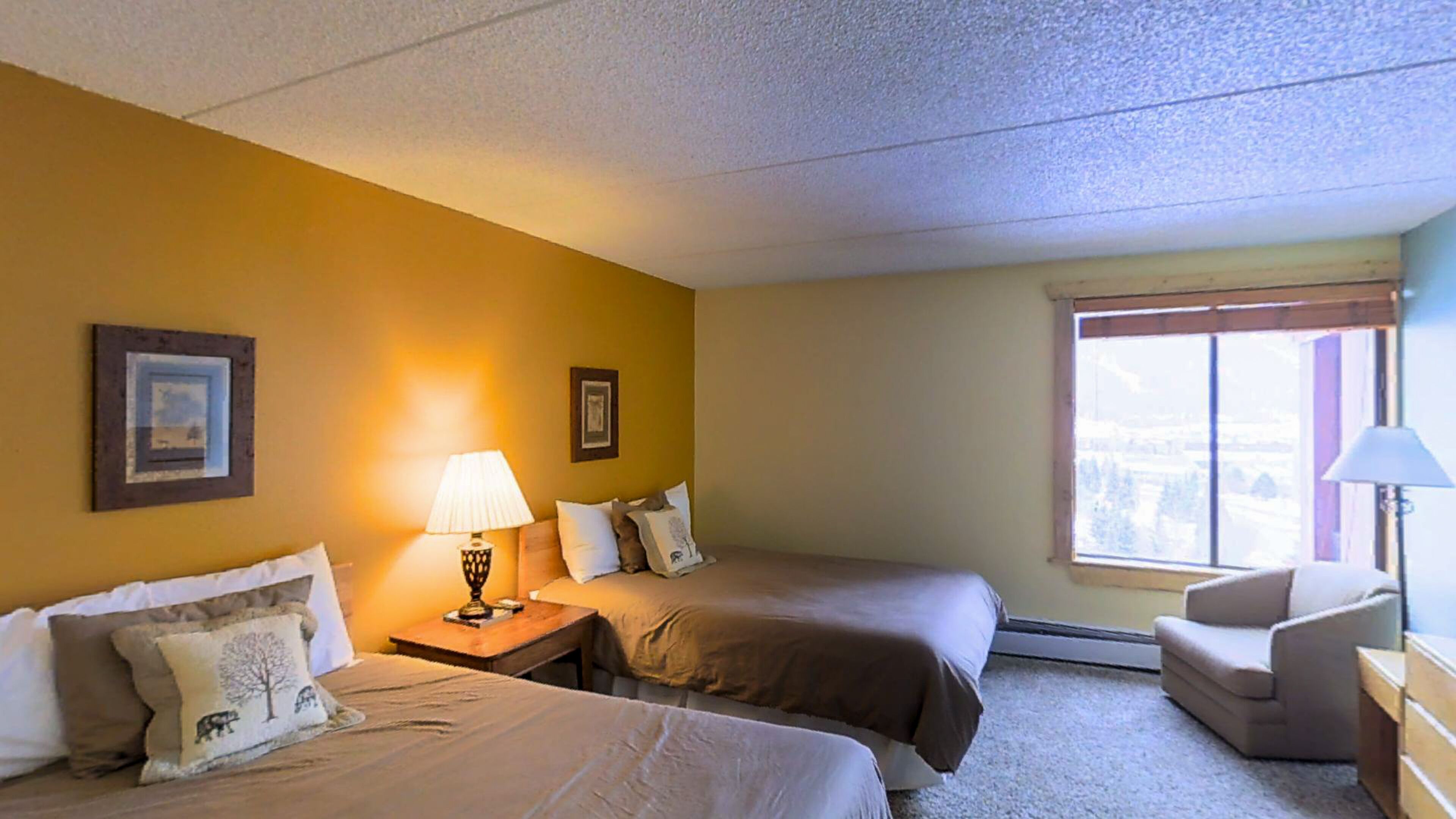 A hotel room with two beds, a nightstand with a lamp, a chair, and a window with a view.