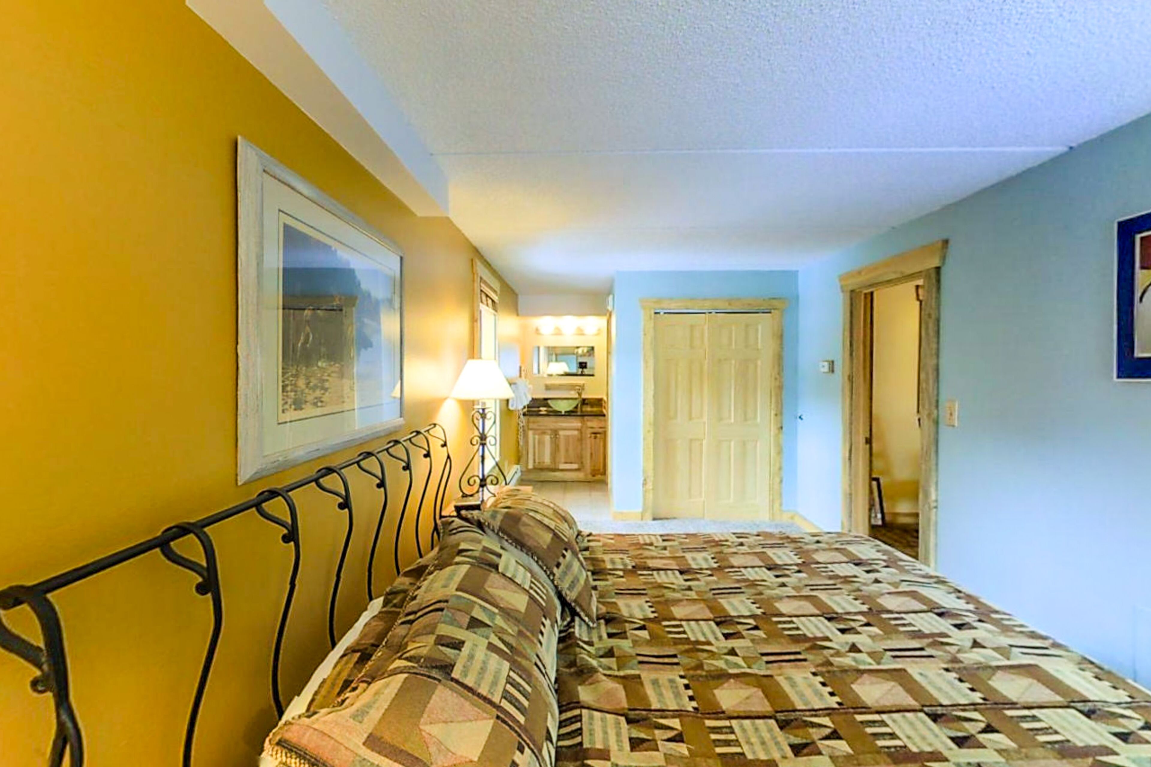A bedroom with a bed, a bedside table, and a lamp. There is a door leading to a bathroom and a hallway.