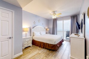 Une chambre à coucher comprenant un lit, des tables de chevet, un ventilateur de plafond, une fenêtre avec des rideaux et une porte.