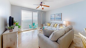 Un salon spacieux avec un canapé gris, un ventilateur de plafond, un téléviseur posé sur un meuble et un balcon agrémenté de chaises vertes.